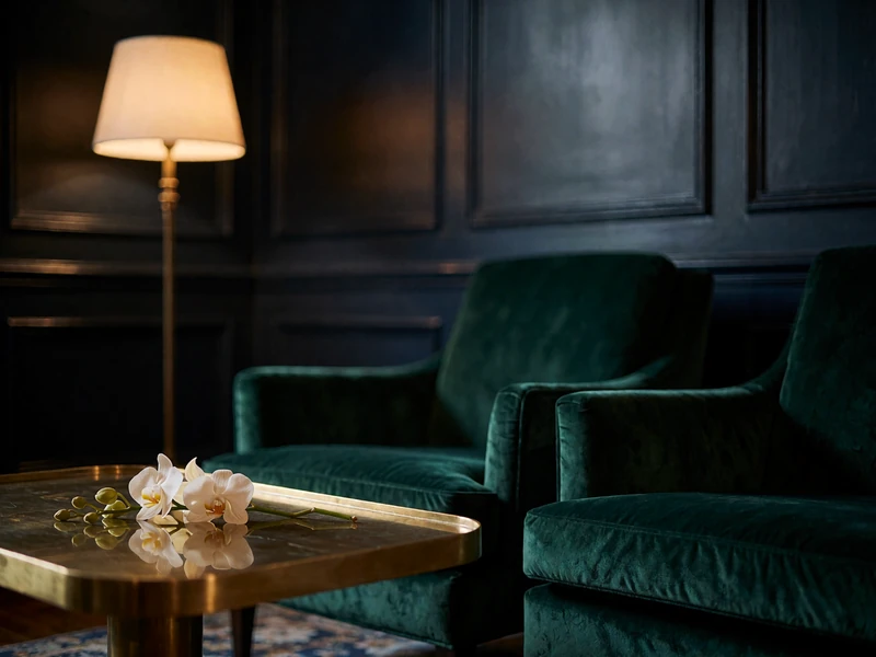 Empty corner of a private members' club lounge with emerald velvet armchairs and a brass-edged table under warm evening light